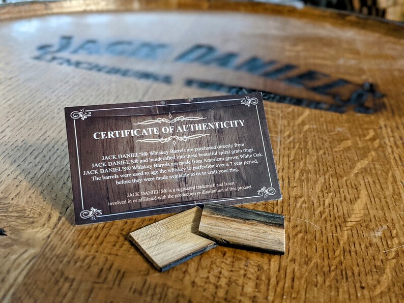 May include: A certificate of authenticity for Jack Daniel's Whiskey Barrel rings. The certificate is printed on a brown card with a gold border. The text on the certificate reads: "Jack Daniel's Whiskey Barrels are purchased directly from Jack Daniel's and handcrafted into these beautiful spiral grain rings. Jack Daniel's Whiskey Barrels are made from American grown White Oak. The barrels were used to age the whiskey to perfection over a 7 year period, before they were made available to us to craft your ring. Jack Daniel's is a registered trademark and is not involved in or affiliated with the production or distribution of this product."