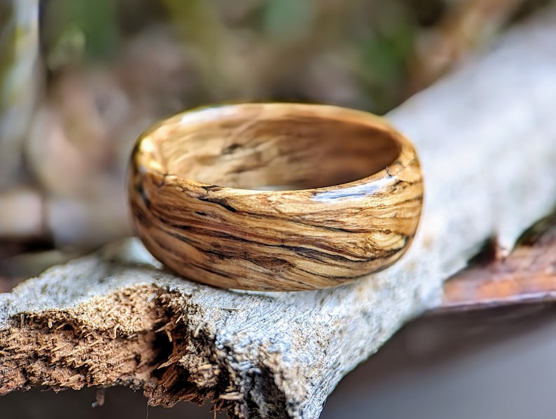 May include: A wooden ring with a natural wood grain pattern and a glossy finish. The ring is brown with dark brown streaks and is resting on a piece of driftwood.