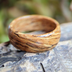 May include: A wooden ring with a natural wood grain pattern. The ring has a dark brown color with light brown streaks throughout. The ring is smooth and polished.