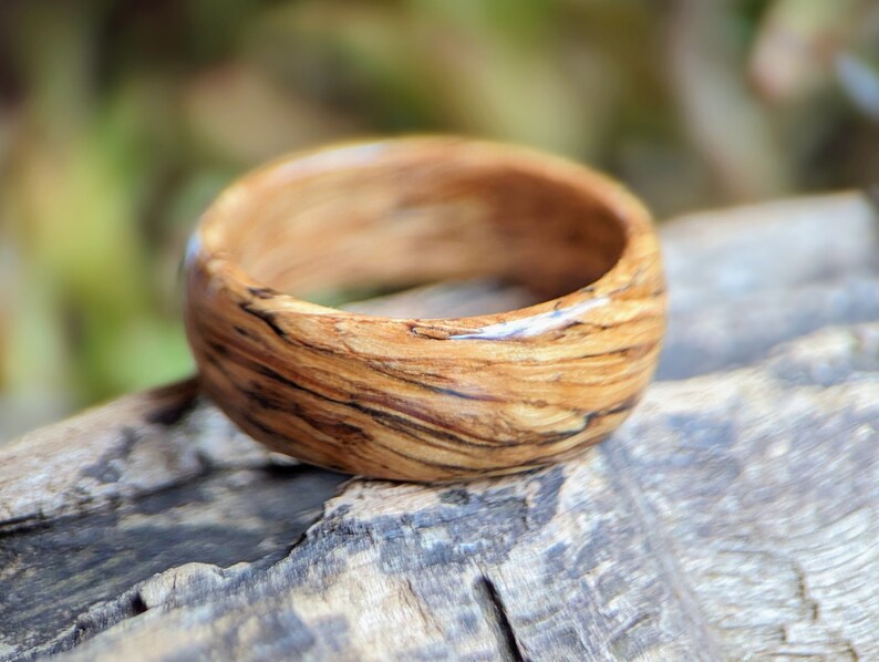 May include: A wooden ring with a natural wood grain pattern. The ring has a dark brown color with light brown streaks throughout. The ring is smooth and polished.