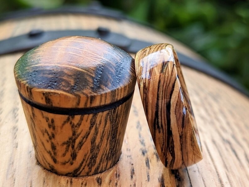 May include: Two wooden rings with a dark wood grain and a clear epoxy finish. The rings are sitting on a wooden surface.