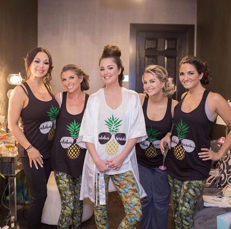 May include: Five women wearing black tank tops with pineapple designs and the words "Aloha" and "Bride" or "Beaches". The bride is wearing a white robe and has her hair up in a bun.