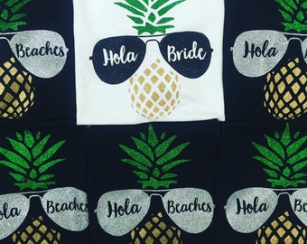 May include: Set of six black and white t-shirts with a pineapple design. The pineapples are wearing sunglasses that say "Hola" and "Beaches". One shirt says "Bride" instead of "Beaches".