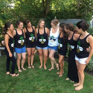 May include: Seven women wearing black tank tops with a pineapple design and the text "Aloha Beaches" printed on them. The women are standing in a group, smiling and looking at the camera.