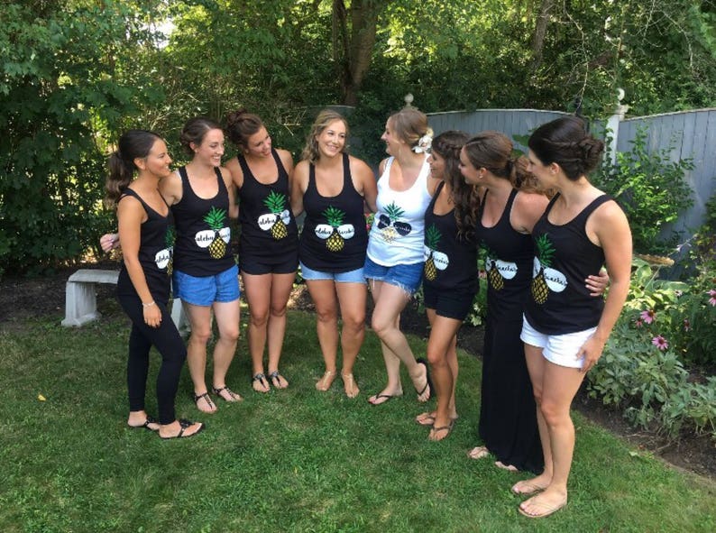 May include: Seven women wearing black tank tops with a pineapple design and the text "Aloha Beaches" printed on them. The women are standing in a group, smiling and looking at the camera.