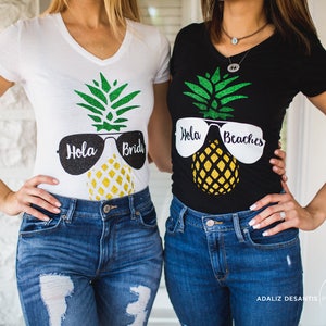 May include: Two women wearing matching shirts with a pineapple design and sunglasses. The shirts say "Hola Bride" and "Hola Beaches".