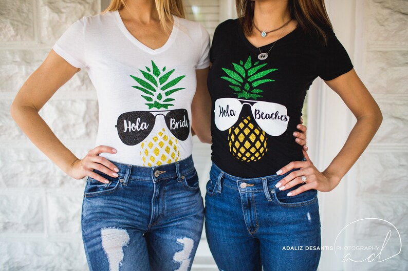 May include: Two women wearing matching shirts with a pineapple design and sunglasses. The shirts say "Hola Bride" and "Hola Beaches".