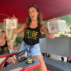 May include: Two women are riding in a party bike. One woman is holding a six-pack of White Claw hard seltzer and the other is holding a single can. The woman holding the six-pack is wearing a black tank top with the text "TRIBE #ScottsdaleBeforeTheVeil" printed on it.