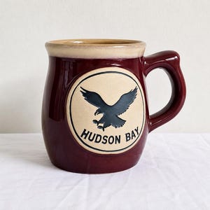 Peut inclure: Une tasse en céramique bordeaux profond avec un bord crème et un emblème circulaire assorti sur le devant. L'emblème présente un aigle bleu foncé en vol au-dessus du texte noir HUDSON BAY, le tout sur un fond beige clair.