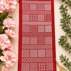 Puede incluir: Una pieza de tela rectangular roja con patrones de bordado geométricos blancos dispuestos en una cuadrícula de cuadrados y bandas horizontales. El bordado consiste en varios diseños de líneas intrincados y repetitivos.