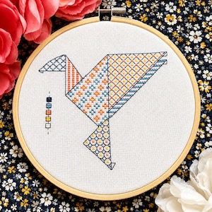 May include: A circular wooden embroidery hoop containing white fabric with a cross-stitched origami bird design. The bird features geometric patterns in blue, orange, and yellow thread. A vertical column of six small colored squares is stitched to the left.
