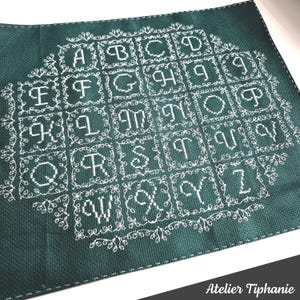 May include: A dark teal cross-stitch sampler featuring the alphabet in white thread. Each letter is framed within a square and surrounded by a decorative floral border. The sampler is stitched on a textured fabric.