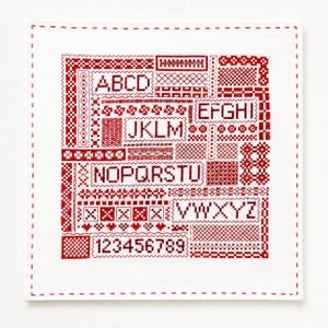以下が含まれることがあります： 赤いクロスステッチ刺繍が施された正方形の白い布製サンプラー。AからZまでのアルファベットと1から9までの数字が、様々な幾何学模様に囲まれた長方形のブロックの中に配置されており、周囲はステッチで縁取られています。