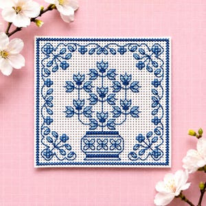 May include: A square cross-stitch embroidery piece featuring a blue floral motif on white Aida cloth. The design depicts a central potted plant with symmetrical branches and leaves, enclosed within a decorative blue geometric border pattern.