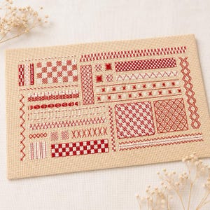 以下が含まれることがあります： ベージュ色の長方形の布地に、赤と白の刺繍による様々なクロスステッチ模様が施されています。刺繍には、幾何学模様、市松模様、ジグザグ、そして整然と並んだX字型のステッチモチーフが含まれています。