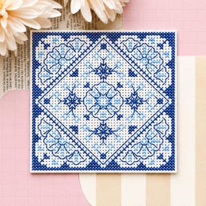 May include: A square cross-stitch embroidery piece featuring a symmetrical geometric pattern in shades of dark blue and light blue on a white fabric grid. The design includes intricate floral-inspired motifs arranged within a central diamond.