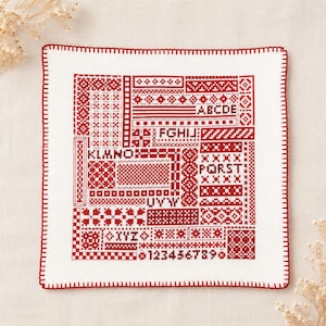 以下が含まれることがあります： 赤いクロスステッチ刺繍が施された正方形の白い布製サンプラー。幾何学模様、AからZまでのアルファベット、1から9までの数字がデザインされています。縁は赤いブランケットステッチで仕上げられています。
