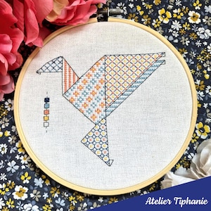 May include: A cross-stitch embroidery of a geometric bird design in a wooden hoop. The bird features orange, blue, and yellow patterns. The text "Atelier Tiphanie" is at the bottom. The background is a floral pattern.