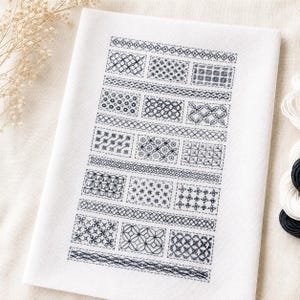 以下が含まれることがあります： 白い布地に黒い刺繍で幾何学模様が施されたサンプラー。長方形のブロックと水平の帯状に配置されており、クロスステッチや線状のデザインなど、さまざまな複雑な刺繍技法が表現されています。