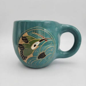 May include: A teal ceramic mug featuring a circular illustration of a hummingbird in flight. The bird is rendered in shades of green, black, and white with gold-coloured outlines. The design includes stylised floral elements and swirling lines.