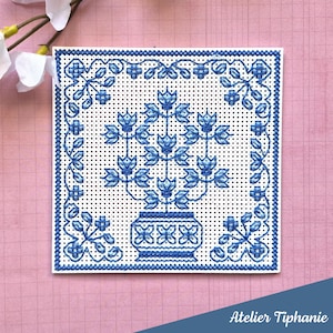 Può includere: Un disegno quadrato a punto croce con un vaso di fiori blu e un bordo su sfondo bianco. Il disegno è ambientato su uno sfondo rosa. Il testo "Atelier Tiphanie" si trova nella parte inferiore dell'immagine.