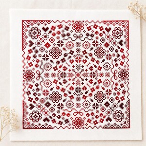 May include: A square piece of white fabric featuring a symmetrical cross-stitch pattern in shades of red and dark maroon. The design consists of intricate geometric shapes, floral motifs, and interconnected lines arranged in a dense, repeating grid.