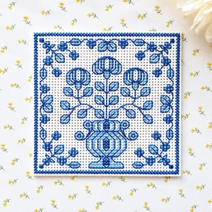 May include: A square cross-stitch embroidery piece featuring a blue and white floral design. The pattern depicts a central vase filled with stylized flowers and leaves, surrounded by a decorative border of small blue geometric shapes.