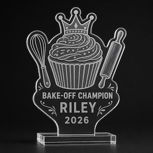 May include: A clear acrylic trophy featuring an etched design of a cupcake topped with a crown, flanked by a whisk and a rolling pin. The text reads BAKE-OFF CHAMPION, RILEY, and 2026. The award stands on a rectangular clear acrylic base.