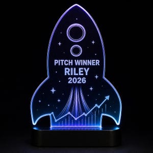May include: A rocket-shaped acrylic LED award plaque with blue and purple edge lighting. The center features the text PITCH WINNER, RILEY, and 2026, surrounded by etched stars, a circular planet, a rising line graph, and rocket exhaust trails.