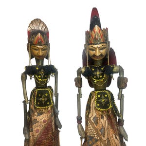 May include: Two Indonesian Wayang Golek puppets. The puppets have carved wooden faces, ornate headdresses, and traditional clothing. The puppets are dressed in black tops with gold accents and patterned skirts. The puppets are on a white background.