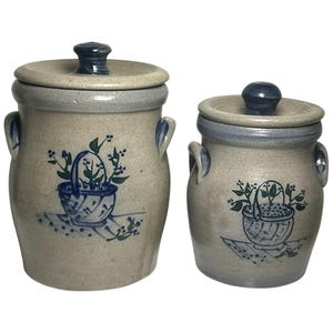 May include: Two beige stoneware canisters with lids, each featuring two side handles and a blue painted design of a basket filled with greenery on a checkered surface. The lids have blue knobs, and the rims are accented with a blue band.
