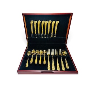 May include: A gold-toned flatware set arranged in a dark wood storage case with a black velvet interior. The set includes eight knives standing upright in the back, with spoons, forks, and a serving spoon laid flat in the front compartments.