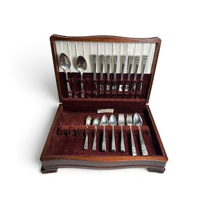 May include: A wooden flatware chest with a hinged lid, featuring a white pleated interior lining and a deep maroon velvet base. The set contains various silver-toned metal spoons, forks, and knives arranged in dedicated slots.