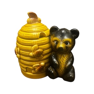 May include: A ceramic honey pot shaped like a beehive with a matching bear figurine. The honey pot is yellow with brown bee and leaf accents. The bear is black and brown.
