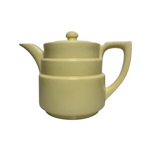 May include: A pale yellow ceramic teapot featuring a stepped, tiered cylindrical body design. The teapot includes a matching lid with a small knob handle, a curved spout on the left side, and a large, angular handle on the right side.