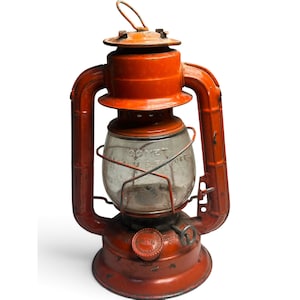 May include: An orange metal kerosene lantern featuring a clear glass globe with the embossed text COMET and MADE IN U.S.A. PAT. OFF. The lantern has a wire handle at the top, a circular base, and a side adjustment knob for the wick mechanism.