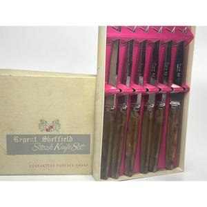 May include: A set of six steak knives with brown marbled handles and stainless steel blades, presented in a beige box. The box has a silver label that reads "Regent Sheffield Steak Knife Set" and "Guaranteed Forever Sharp".