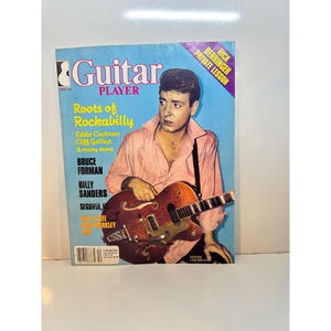 May include: A vintage Guitar Player magazine featuring a man holding a brown guitar. The cover has a blue background with the text "Guitar Player" and "Roots of Rockabilly". The magazine also features the text "Eddie Cochran" and "Rick Derringer Private Lesson".