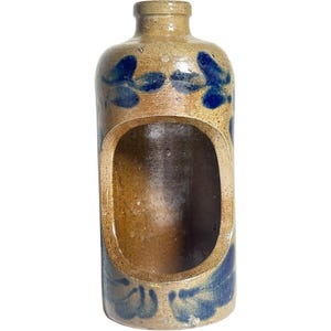 May include: A tan stoneware bottle with a narrow neck and a large, oval-shaped cutout on the front. The exterior features hand-painted cobalt blue floral motifs around the top and bottom sections, with a smooth, glazed finish throughout.