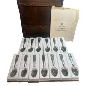 May include: A collection of silver spoons, each in a white foam insert, with a wooden spoon rack and a booklet. The spoons feature detailed engravings. The booklet reads "The Official Bicentennial Spoon Collection."