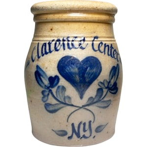May include: A beige stoneware crock with a thick rim and a cylindrical body. The front features blue hand-painted text reading Clarence Center, a central heart motif, floral sprigs, and the letters NY at the bottom of the vessel.