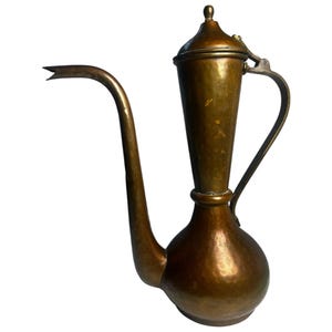 May include: A tall, hammered brass ewer with a bulbous base, a slender neck, and a long, curved spout. The vessel features a hinged lid with a small finial at the top and a curved handle attached to the side, showcasing a weathered, metallic finish.