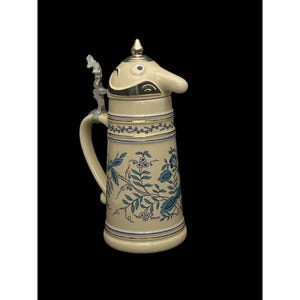 May include: Cream-colored ceramic beer stein with a whimsical, nose-shaped lid and a silver-toned handle. The body features blue floral patterns and gold trim. A decorative piece for beer enthusiasts or collectors.