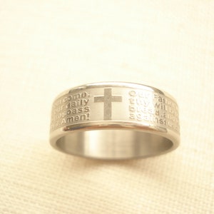 Puede incluir: Un anillo plateado con texto grabado y un diseño de cruz. El anillo presenta el Padre Nuestro, con las palabras grabadas alrededor de la banda. La cruz está centrada en la superficie del anillo.