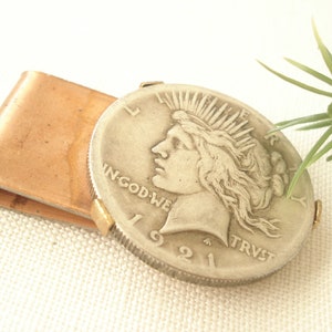 May include: A copper money clip with a silver 1921 Peace Dollar coin attached. The coin features a profile of a woman with flowing hair and the words "IN GOD WE TRUST" and "LIBERTY".