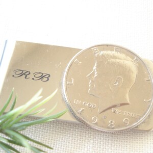 May include: A silver money clip with a half dollar coin featuring John F. Kennedy's profile. The coin has the inscription "In God We Trust" and the year "1969". The money clip is engraved with the initials "R.B."