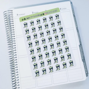 May include: A sheet of stickers featuring a repeating pattern of cartoon green dragons wearing sunglasses and holding a cup. The stickers are on a white background and have the text "SweetttDesigns" and "@sweetttdesigns" printed on them.