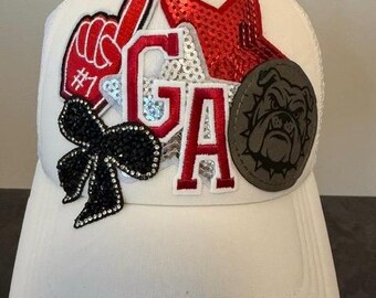 Può includere: Un cappellino da baseball bianco con diverse toppe applicate sulla parte anteriore. Le decorazioni includono un dito in gommapiuma rosso con il numero 1, un fiocco di cristalli neri, stelle di paillettes rosse e argento, le lettere GA e un bulldog.
