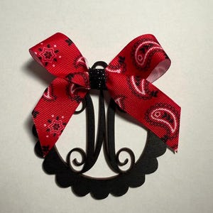 Pu&ograve; includere: Un monogramma nero con un fiocco rosso paisley. Il monogramma ha la forma della lettera "M" ed &egrave; circondato da un bordo smerlato. Il fiocco &egrave; realizzato in nastro rosso con un motivo paisley bianco.