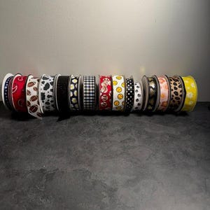 Pu&ograve; includere: Una collezione di rocchetti di nastro decorativo con vari motivi, tra cui paisley, chicchi di caff&egrave;, stampa mucca, margherite, quadretti vichy, palline da baseball e impronte di zampe. I nastri sono in vari colori, come rosso, nero, bianco e giallo.
